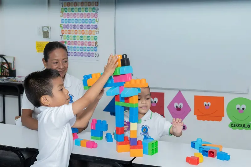 Una maestra acompaña a dos alumnos de preescolar que construyen una torre con bloques de colores